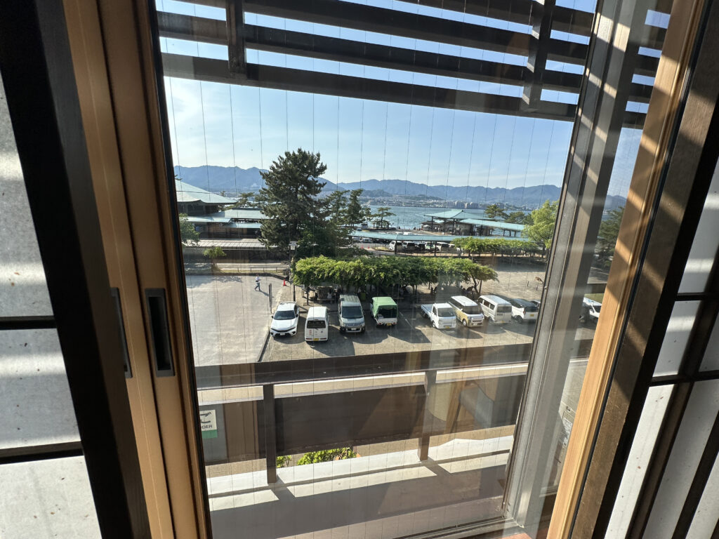 The image shows a view through a window, looking out onto a parking lot with several cars parked. Beyond the parking lot, there are trees and a body of water, possibly a lake or sea, with mountains visible in the distance. The sky is clear and sunny. The window has vertical blinds, and the interior appears to be part of a building with wooden frames.