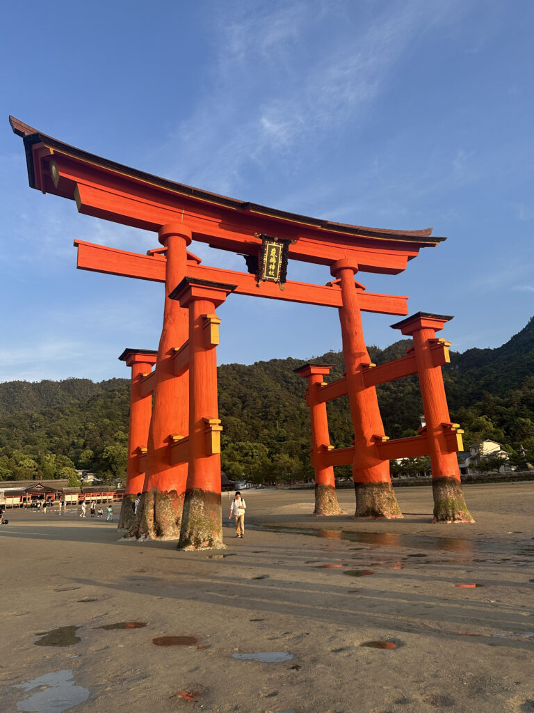 A large, traditional Japanese torii gate stands prominently on a sandy beach with a backdrop of green hills and a clear blue sky. The gate is painted bright orange with black and gold accents. People are walking around the area, and there are small puddles on the sand, suggesting a recent low tide.