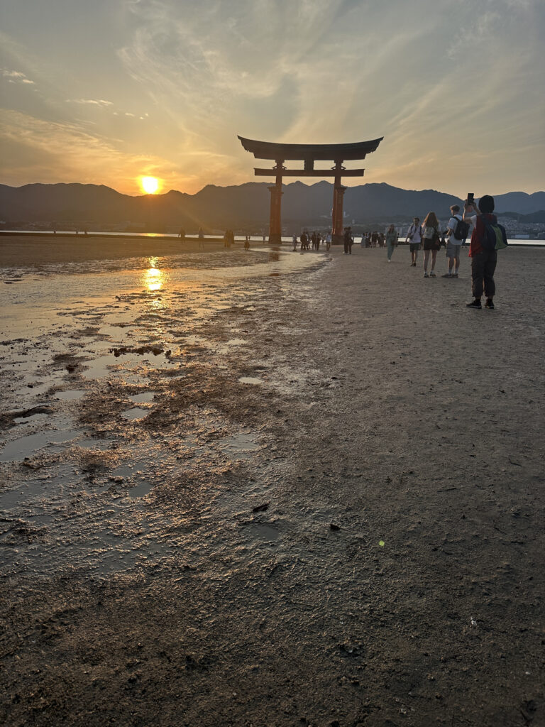 The image shows a scenic view of a large torii gate standing in the water during low tide, with a group of people walking towards it. The sun is setting behind the mountains in the background, casting a warm glow over the scene. The foreground features wet sand with reflections of the sunlight.