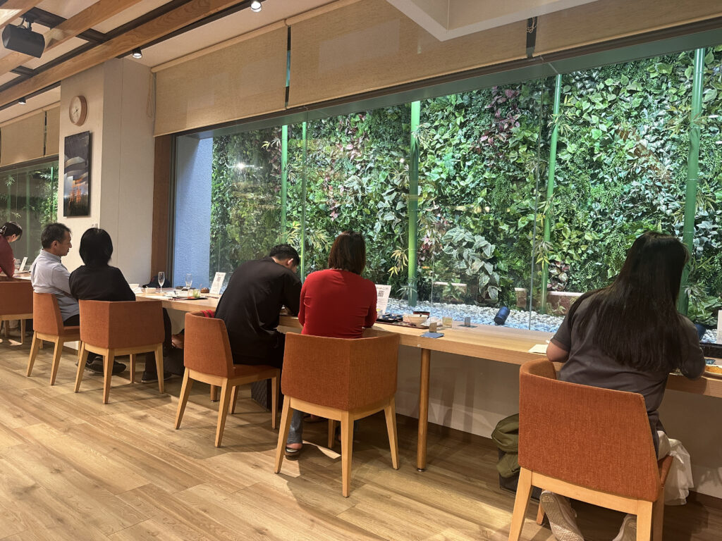 The image shows a group of people sitting at a long wooden table in a modern indoor setting. They are seated on orange chairs, facing a large window with a view of a lush vertical garden filled with various green plants. The room has wooden flooring and a ceiling with exposed beams. A clock and a framed picture are visible on the wall. The atmosphere appears to be calm and relaxed.