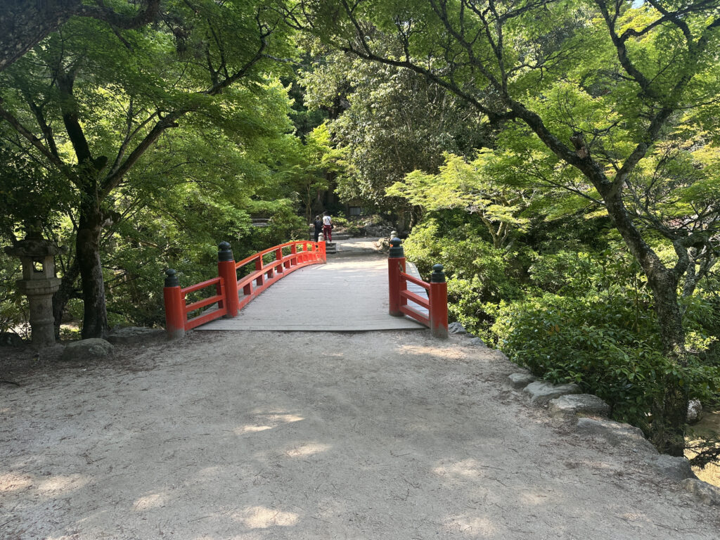 A serene scene featuring a traditional red wooden bridge with black accents, surrounded by lush green trees. The bridge crosses over a small stream or path, and a person is visible in the distance walking across it. The area is shaded by the dense foliage, creating a peaceful, natural atmosphere.