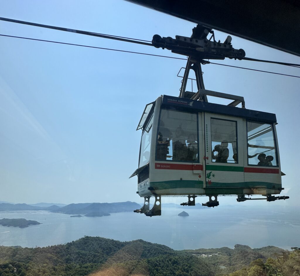 A cable car is suspended in the air, carrying passengers. It is part of the Miyajima Ropeway, as indicated by the text on the car. The background features a scenic view of the ocean and distant islands under a clear blue sky. The landscape below includes lush green hills.