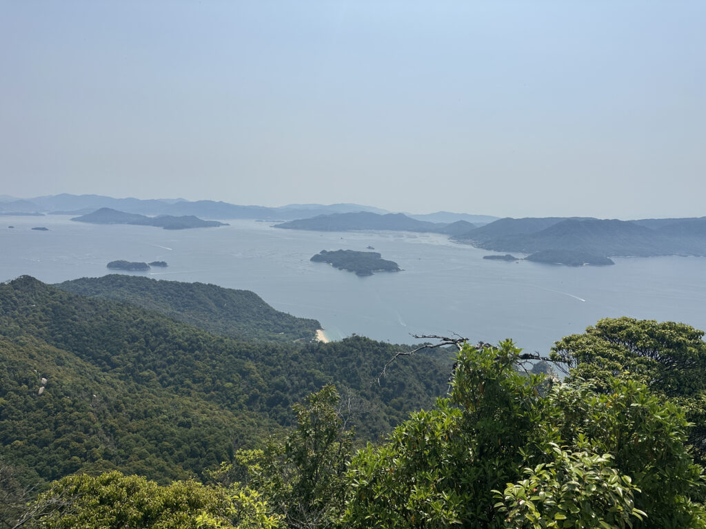 A scenic view of a coastal landscape featuring lush green hills in the foreground and a vast expanse of water dotted with small islands. The horizon shows a range of distant mountains under a clear blue sky.