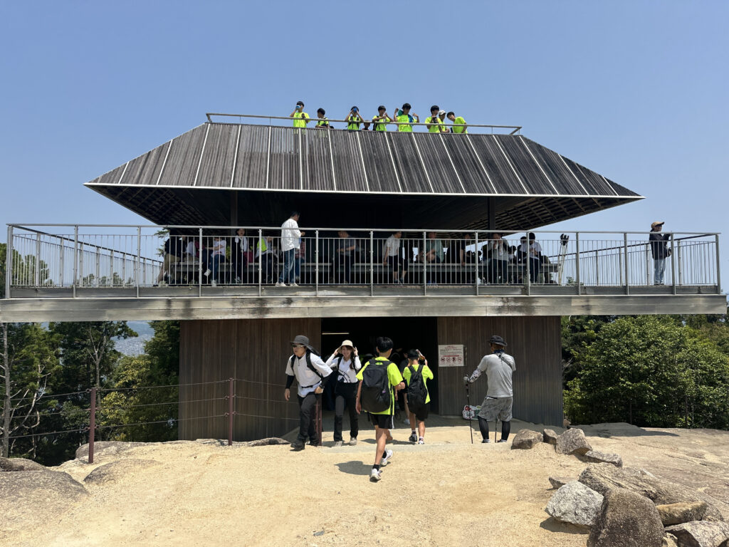 A modern, angular building with a sloped roof and two levels of observation decks. People are gathered on both levels, with some wearing bright yellow vests. The structure is elevated, surrounded by trees, and set against a clear blue sky. A few individuals are walking towards the building on a sandy path.