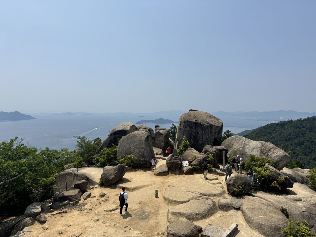 A scenic view from a rocky hilltop overlooking a vast body of water with several small islands in the distance. The foreground features large boulders and a few people walking and exploring the area. The background shows a clear blue sky and distant mountains.