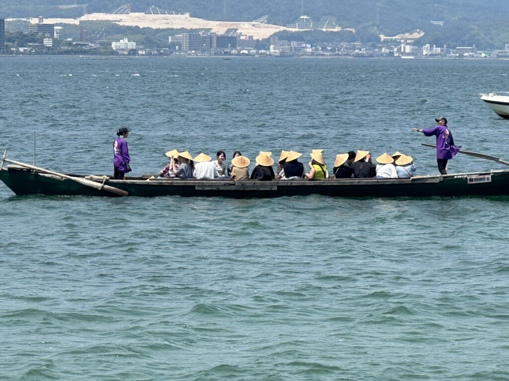 A long wooden boat is on the water, carrying a group of people. Most of the passengers are wearing traditional straw hats. Two individuals in purple clothing are standing at either end of the boat, possibly guiding or steering it. The background shows a distant shoreline with buildings and hills.