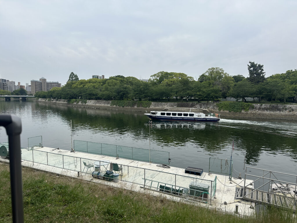 The image shows a river with a boat traveling along it. The riverbank is lined with green trees and there are buildings visible in the background. In the foreground, there is a dock with some equipment and chairs. The sky is overcast.