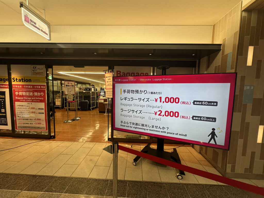 The image shows the entrance to a "Luggage Station" at a location identified as Shin-Osaka. There is a large digital sign displaying information about baggage storage services. The sign lists prices for regular and large baggage storage, with regular size costing ¥1,000 and large size costing ¥2,000. The text is in both Japanese and English. Inside the station, there are luggage racks and storage areas visible. The floor is wooden, and there are signs and posters on the walls.