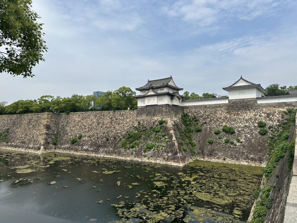 The image shows a traditional Japanese castle wall with a moat in the foreground. The stone wall is topped with white structures featuring traditional Japanese architectural elements, such as tiled roofs. The moat contains water with patches of green algae or aquatic plants. Trees and greenery are visible in the background under a partly cloudy sky.