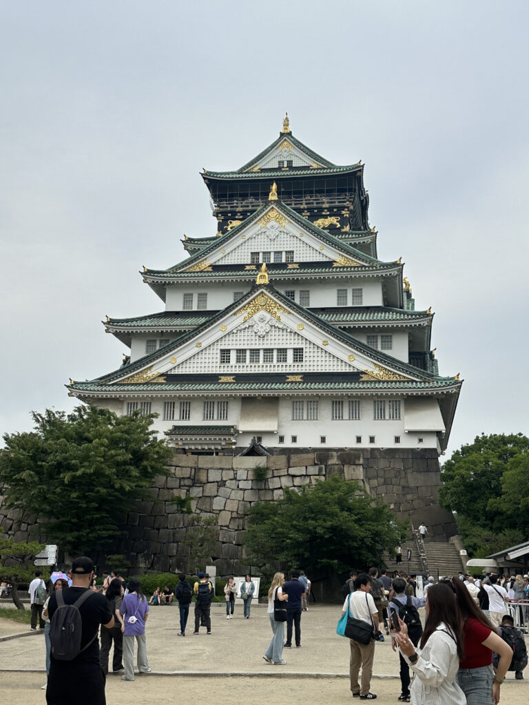 The image shows Osaka Castle, a historic Japanese castle with multiple tiers and ornate architectural details, including green roofs and gold accents. It is surrounded by stone walls and trees. In the foreground, there are many people walking and taking photos, indicating it is a popular tourist destination. The sky is overcast.