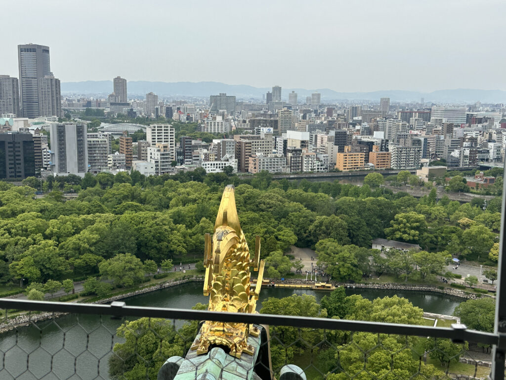 A cityscape view from a high vantage point, showing a mix of modern buildings and lush green trees. In the foreground, there is a golden ornamental structure, possibly part of a historic building or castle. A river or moat runs through the greenery, and a boat is visible on the water. The sky is overcast, and the city extends into the distance with mountains on the horizon.