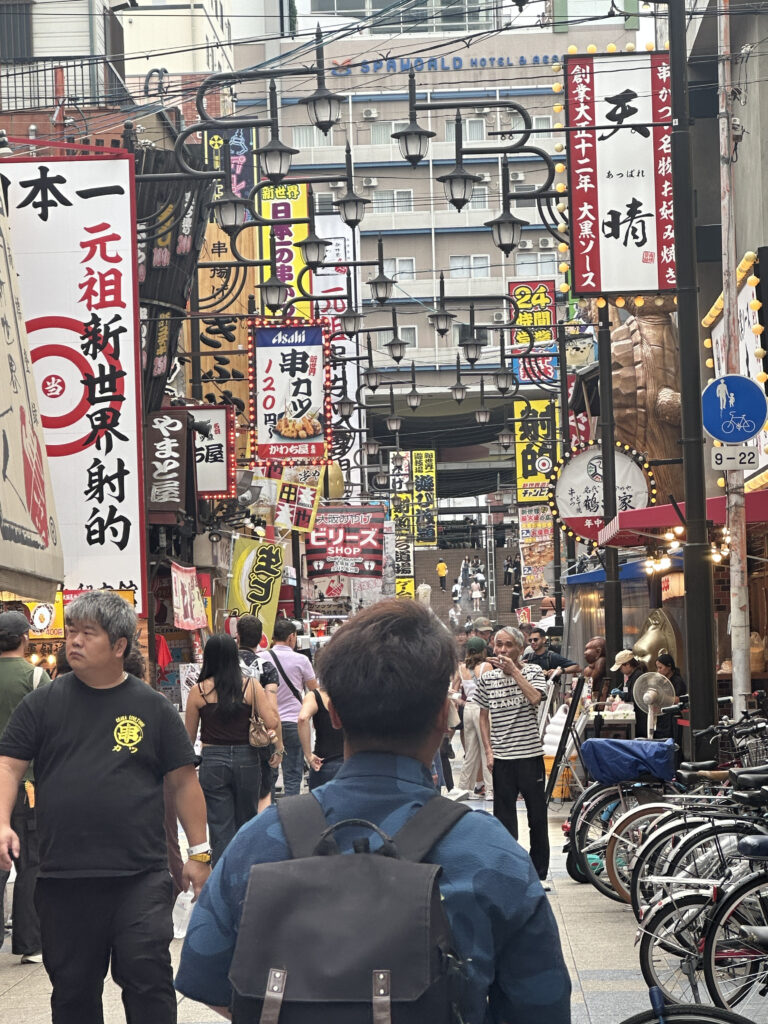 A bustling street scene in a Japanese city, filled with people walking and shopping. The street is lined with numerous colorful signs and banners featuring Japanese text. There are bicycles parked on the right side, and various shops and eateries are visible. The atmosphere is lively and vibrant, typical of a busy urban area.