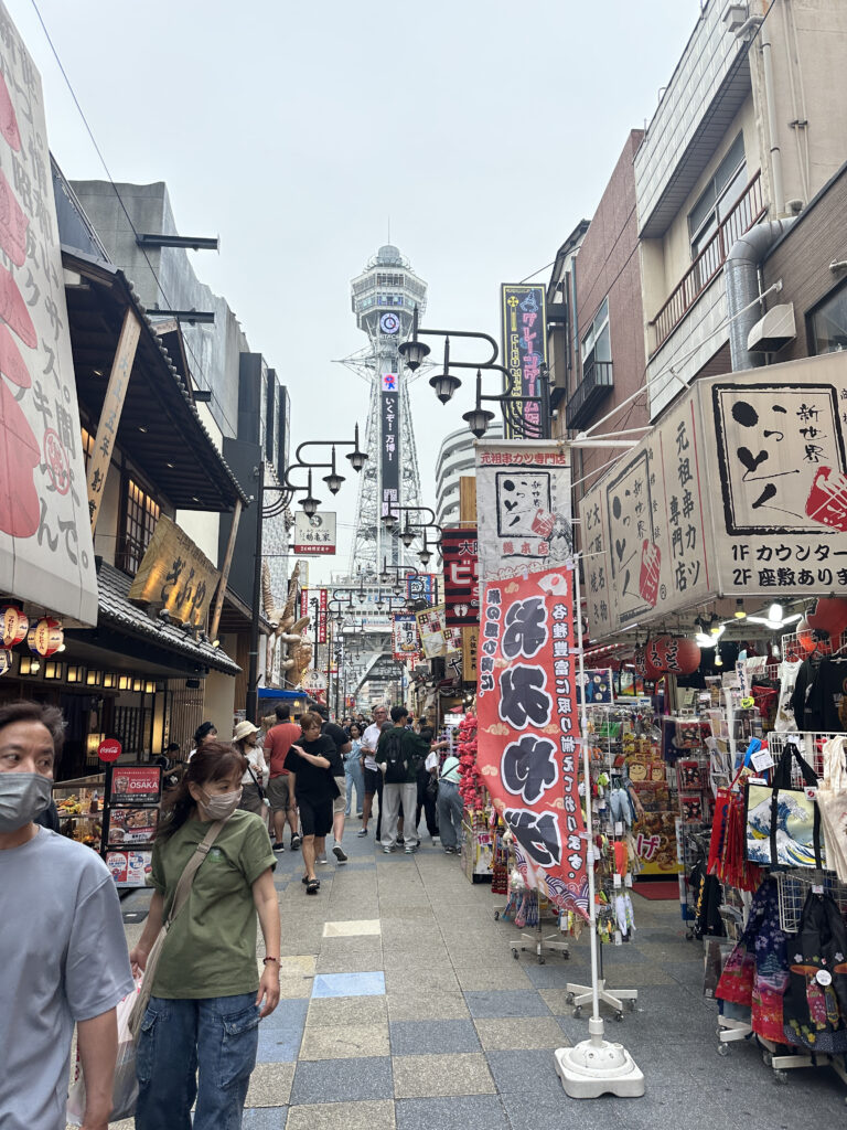A bustling street scene in a Japanese shopping district. The street is lined with various shops displaying colorful signs and banners with Japanese text. People are walking along the street, some wearing masks. In the background, a tall tower is visible, rising above the buildings. The atmosphere is lively and vibrant, typical of a busy urban area.