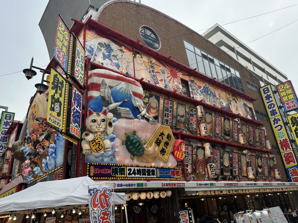 The image shows a colorful and vibrant building facade in a busy urban area, likely in Japan. The building is adorned with large, eye-catching signs and decorations, including a giant maneki-neko (beckoning cat), sumo wrestler illustrations, and various Japanese characters. The signs are brightly colored with reds, yellows, and blues, and there are lanterns and other decorative elements. The building appears to be a restaurant or entertainment venue, with a tent and lights in front of it.