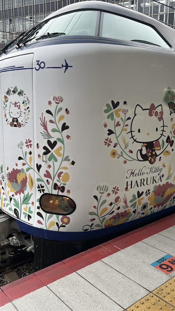 The image shows the front of a train decorated with Hello Kitty-themed artwork. The design features Hello Kitty in a kimono surrounded by colorful floral patterns. The words "Hello Kitty HARUKA" are also visible on the train. The train is at a station platform.