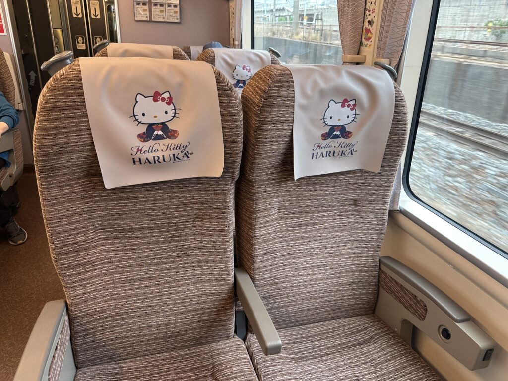 The image shows the interior of a train with two seats covered in brown fabric. Each seat has a white headrest cover featuring a Hello Kitty design and the text "Hello Kitty HARUKA." The train window on the right side reveals a view of the tracks and the outside scenery.