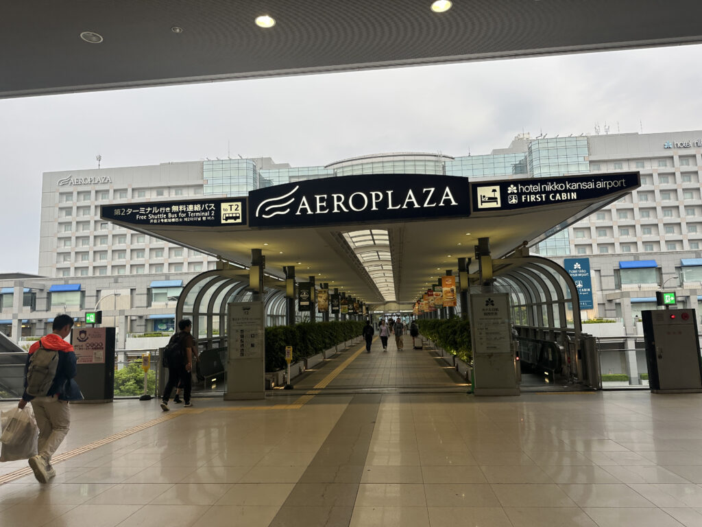 The image shows a covered walkway leading to a building labeled "AEROPLAZA." Above the walkway, there are signs indicating directions to a free shuttle bus for Terminal 2 and accommodations at Hotel Nikko Kansai Airport and First Cabin. People are walking along the path, and the background features a large building with multiple windows. The area appears to be part of an airport complex.