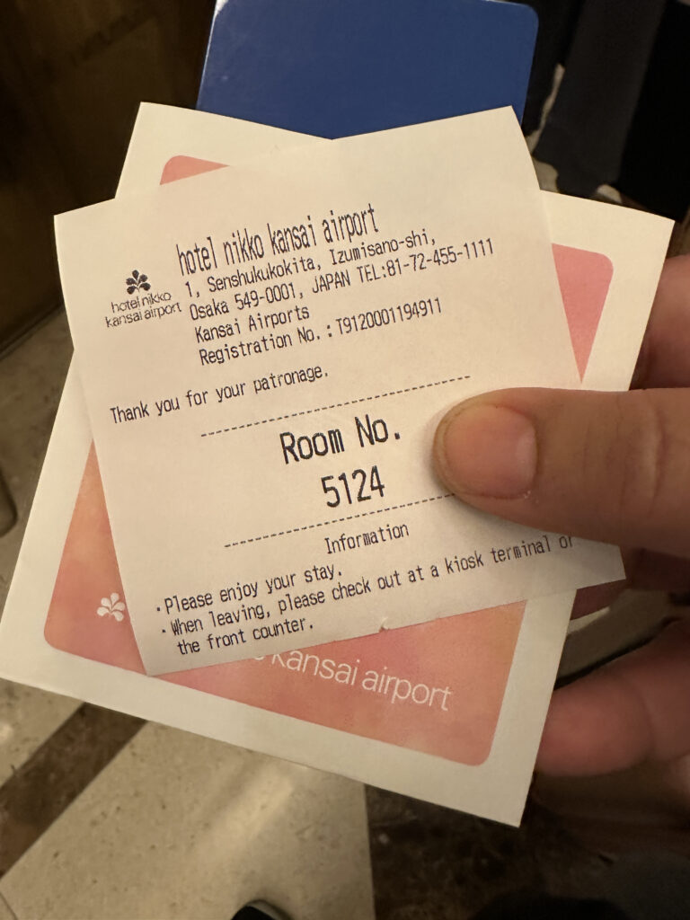 The image shows a hand holding a hotel key card and a paper slip from Hotel Nikko Kansai Airport. The slip includes the hotel's address in Osaka, Japan, and a phone number. It also displays the room number, 5124, and provides information about checking out. The background shows a marble floor.