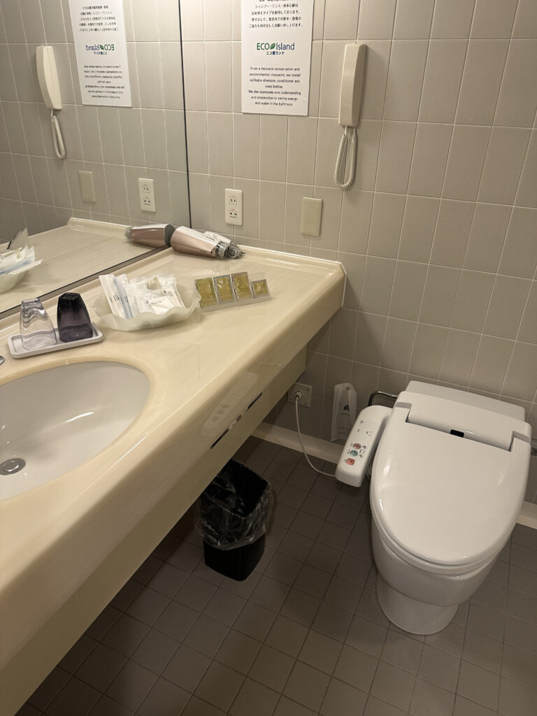 The image shows a bathroom with a modern toilet featuring a bidet control panel on the side. To the left, there is a countertop with a sink. On the countertop, there are various toiletries, including a hairdryer, wrapped items, and small bottles. Above the sink is a mirror with informational signs in both English and Japanese. The floor is tiled, and there is a small trash bin under the counter.