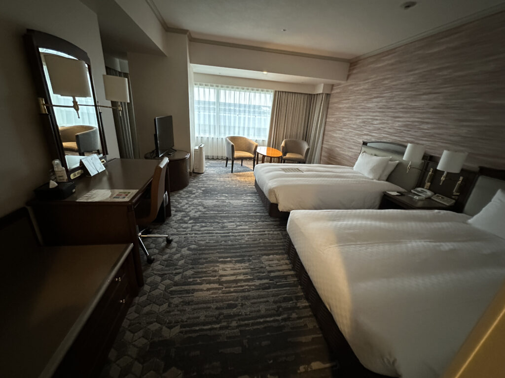 The image shows a hotel room with two neatly made twin beds. There is a nightstand between the beds with lamps and a telephone. A desk with a chair and a mirror is on the left side, and a flat-screen TV is placed on a cabinet. Near the window, there are two armchairs and a small round table. The room has a patterned carpet and a large window with sheer curtains, allowing natural light to enter.