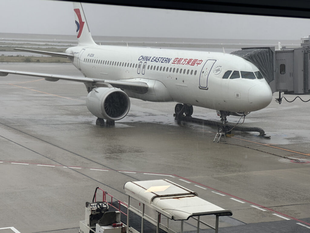 The image shows a China Eastern Airlines aircraft parked at an airport gate. The plane is connected to a jet bridge, and the ground is wet, indicating rainy weather. The aircraft is white with the airline's logo and name visible on the fuselage. A ground service vehicle is seen in the foreground.