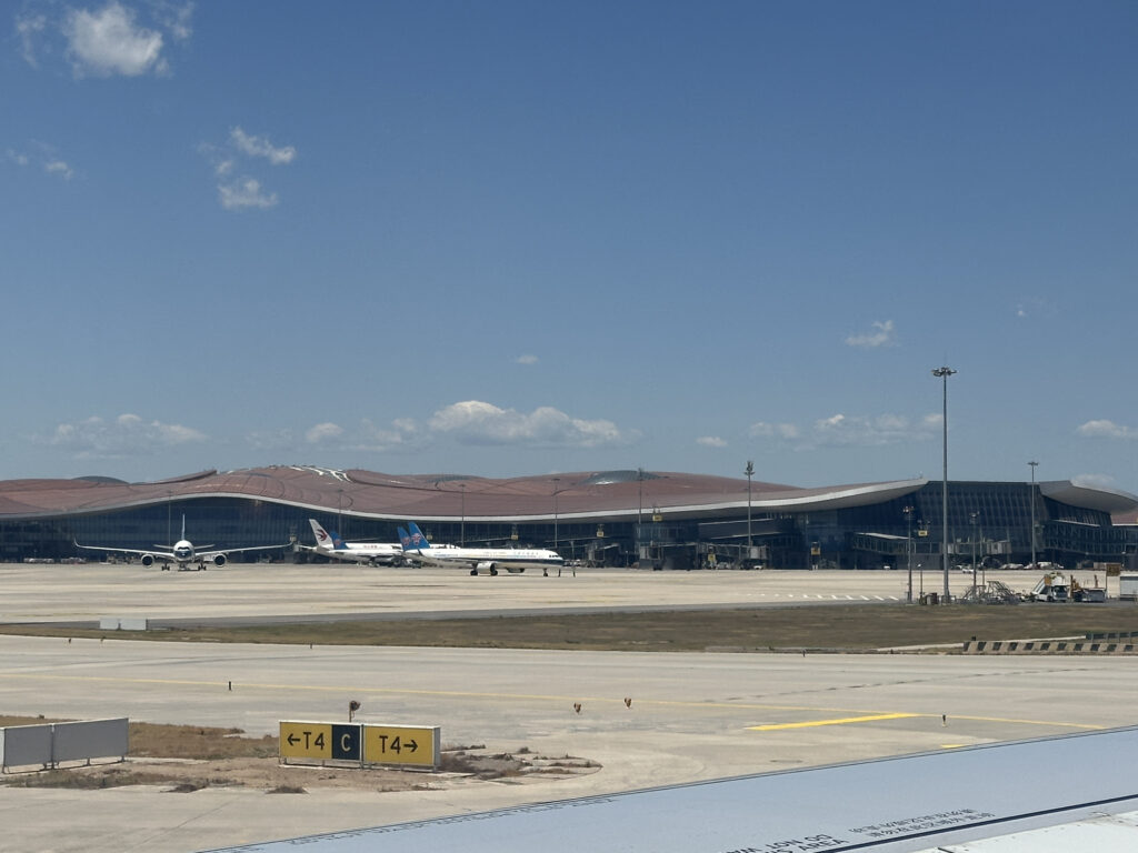 The image shows an airport terminal with a modern, wavy roof design. Several airplanes are parked on the tarmac in front of the terminal. The sky is clear with a few clouds, and there are signs on the ground indicating directions for taxiing aircraft.