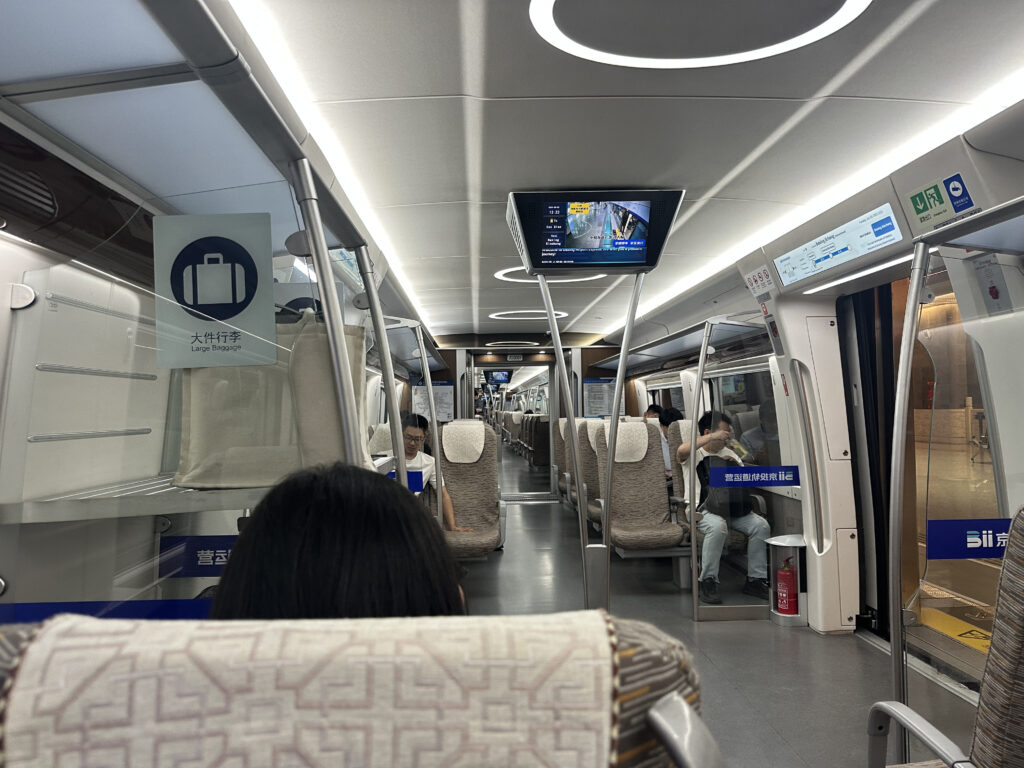 The image shows the interior of a modern train carriage. The seating is arranged in rows with beige and brown patterned upholstery. Overhead, there are luggage racks with a sign indicating "Large Baggage" in both English and Chinese. A digital screen is mounted on the ceiling, displaying information. Passengers are seated, some looking at their phones. The train has a clean and well-lit environment, with emergency instructions and signs visible near the doors.