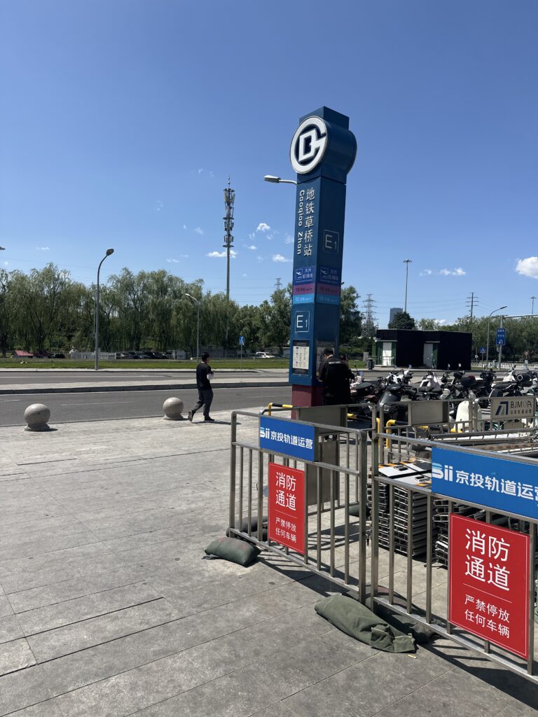 The image shows an outdoor scene with a tall signpost for a subway station labeled "E1" in Chinese characters. There are barriers with signs in Chinese, and a few sandbags at the base. In the background, there are trees, a road, and a person walking. The sky is clear and blue.