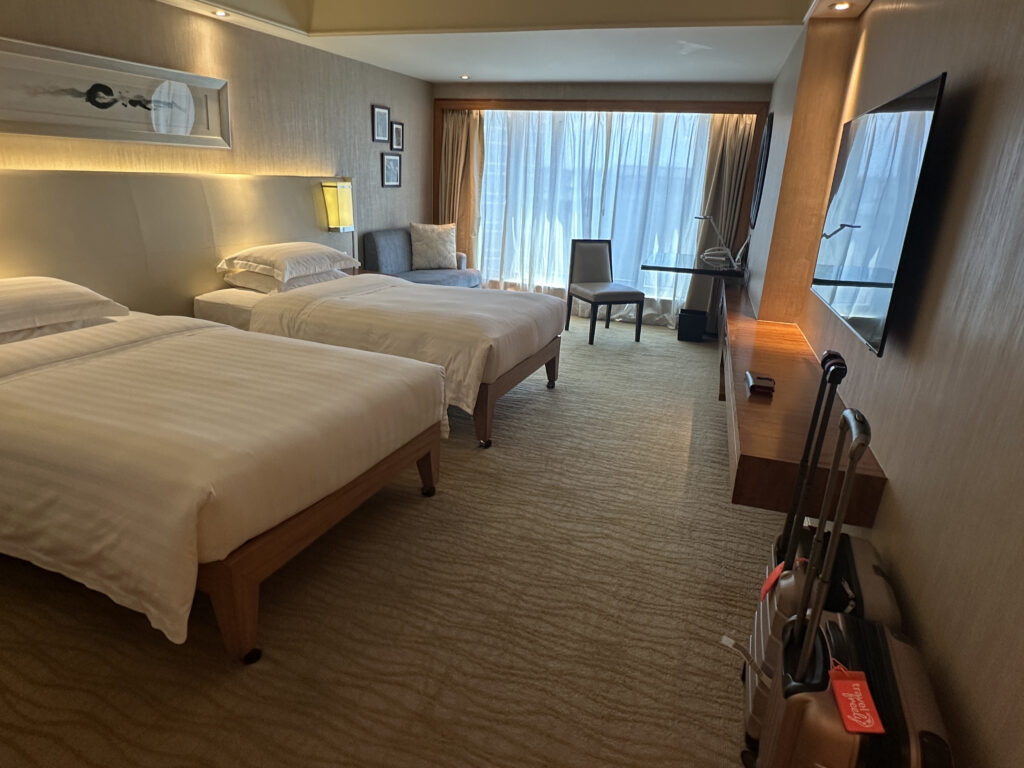 The image shows a modern hotel room with two neatly made single beds. The room features a large window with sheer curtains, allowing natural light to fill the space. There is a chair and a small table near the window. On the opposite wall, a flat-screen TV is mounted above a long wooden desk. Two suitcases with red tags are placed near the desk. The room has a neutral color palette with beige walls and carpet.