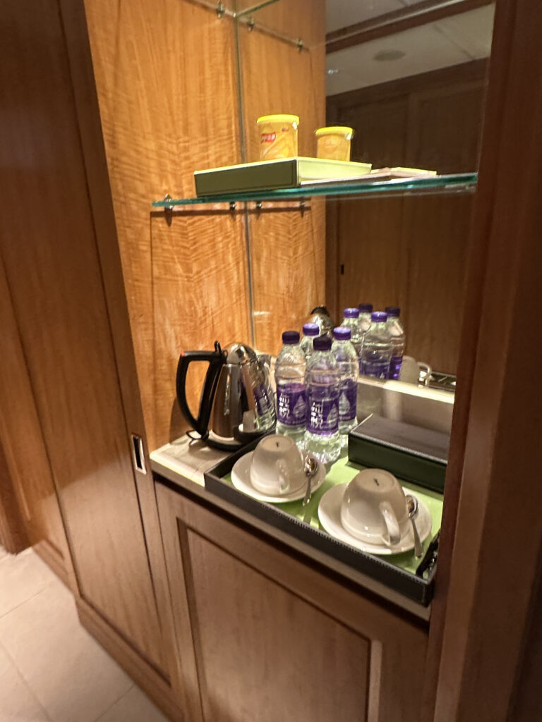 The image shows a hotel room refreshment station. It includes a wooden cabinet with a glass shelf. On the shelf, there are two containers with yellow lids. Below, there is a tray with a kettle, several bottles of water, and two cups with saucers and spoons.