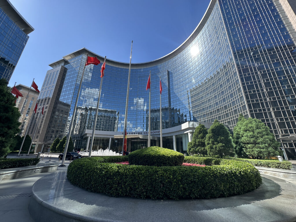 The image shows a modern, curved glass building with a reflective facade, surrounded by well-maintained landscaping. Several red flags are flying on flagpoles in front of the building. There are neatly trimmed bushes and trees in the foreground, and a fountain is visible near the entrance. The sky is clear and blue, with a few clouds.