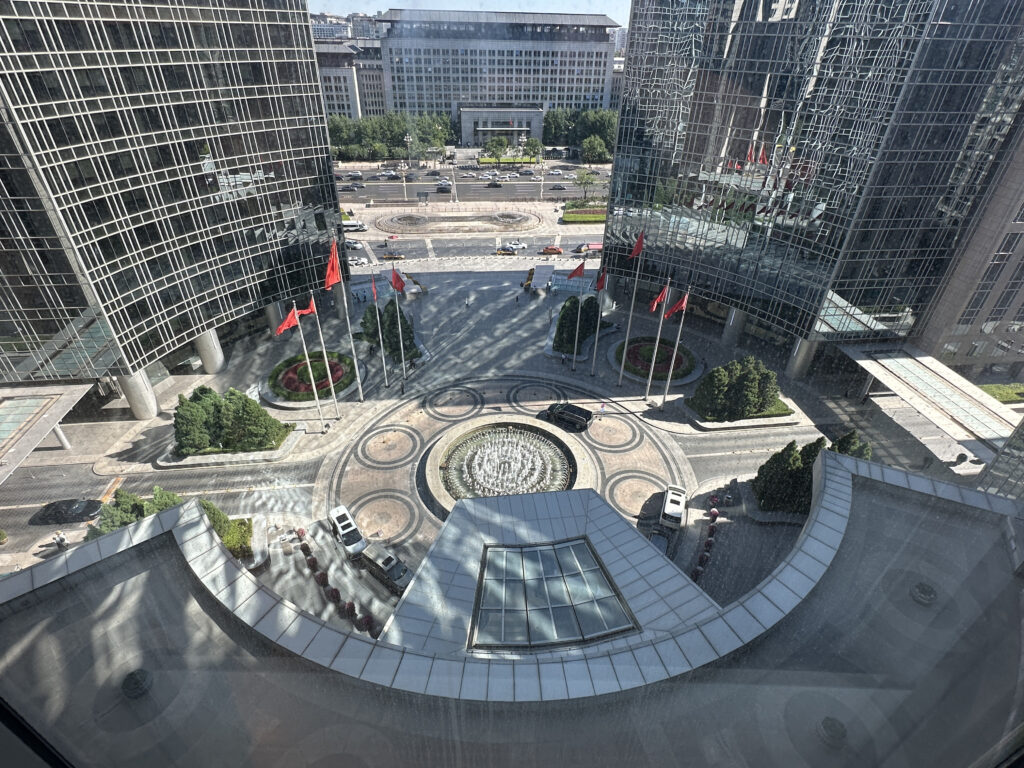 The image shows a view from a high vantage point overlooking a modern urban area. There are two tall glass buildings with reflective surfaces, and a circular plaza below featuring a fountain in the center. Several red flags are displayed on flagpoles around the plaza. The area is landscaped with trees and shrubs, and there are a few vehicles parked near the buildings. In the background, more buildings and a road are visible.