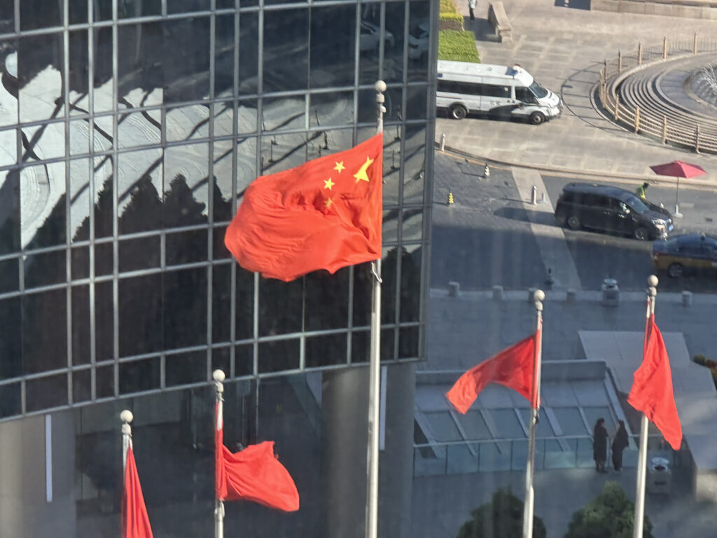 The image shows several red flags with yellow stars, resembling the flag of China, waving in the wind. They are mounted on flagpoles in front of a modern glass building. In the background, there is a street with a white van and a black car parked nearby. The area appears to be an urban setting with a paved plaza and some people walking.