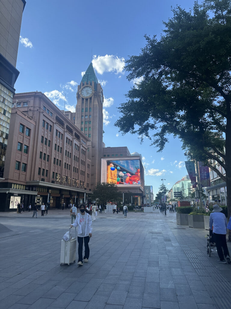 A city street scene with a tall building featuring a clock tower on the left. The building has a pinkish hue and a green roof. There is a large digital billboard displaying colorful advertisements. People are walking along the wide pedestrian area, and a person in the foreground is pulling a suitcase. Trees line the right side of the street, and the sky is clear with a few clouds.