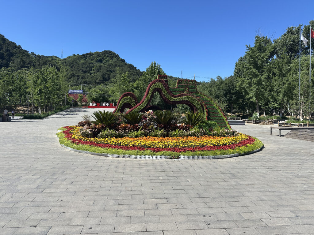 A landscaped garden with a large, decorative flower arrangement in the center, featuring a wavy, arch-like structure made of greenery and flowers. The garden is surrounded by a paved area, with trees and hills in the background under a clear blue sky.