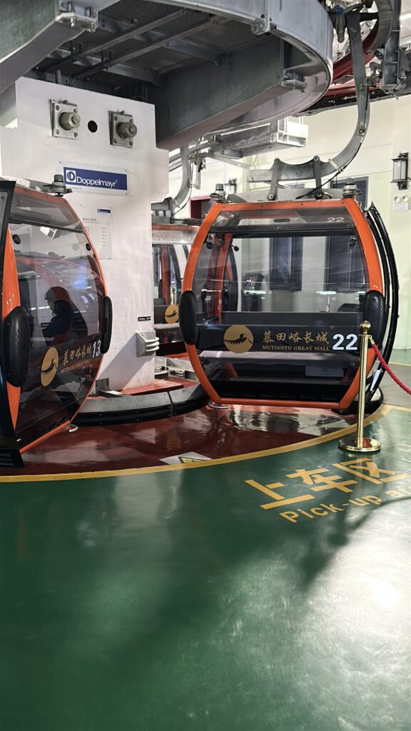 The image shows a cable car station with two gondolas labeled "Mutianyu Great Wall." The gondolas are orange and black, with transparent windows, and are numbered 13 and 22. The station has a green floor with yellow text indicating a "Pick-up area." A red rope barrier is visible near the gondolas. The brand "Doppelmayr" is displayed on a sign above.