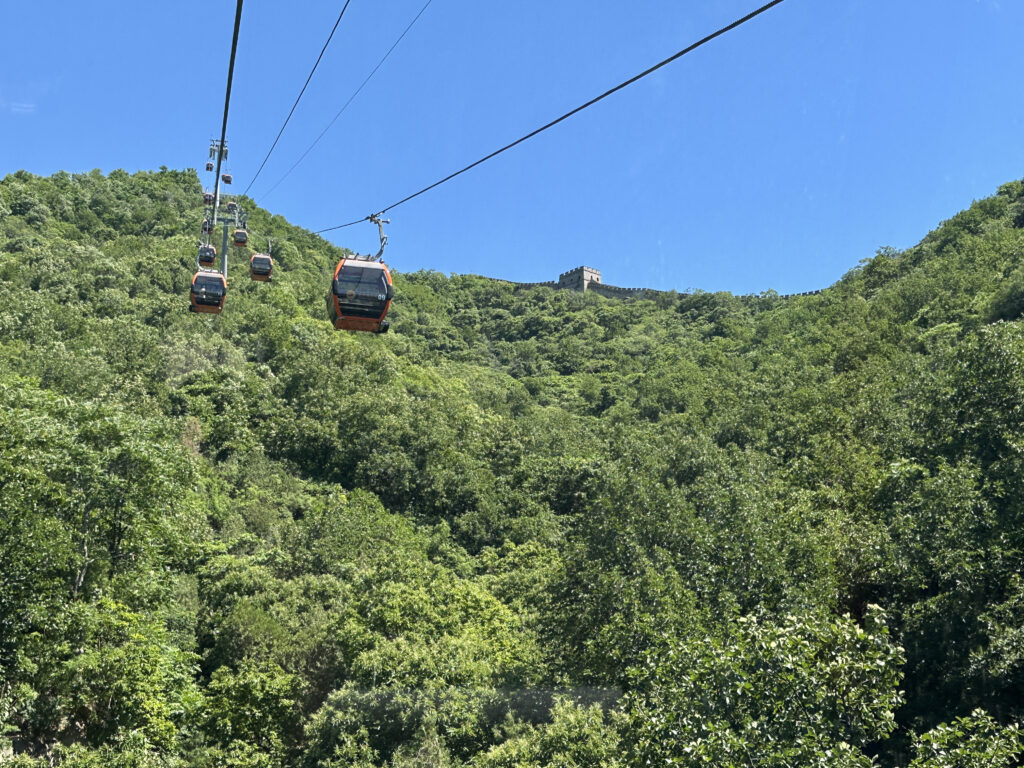 The image shows a series of cable cars traveling over a lush, green forested hillside. In the background, a section of a historic stone wall is visible, likely part of the Great Wall of China. The sky is clear and blue, providing a bright backdrop to the scene.