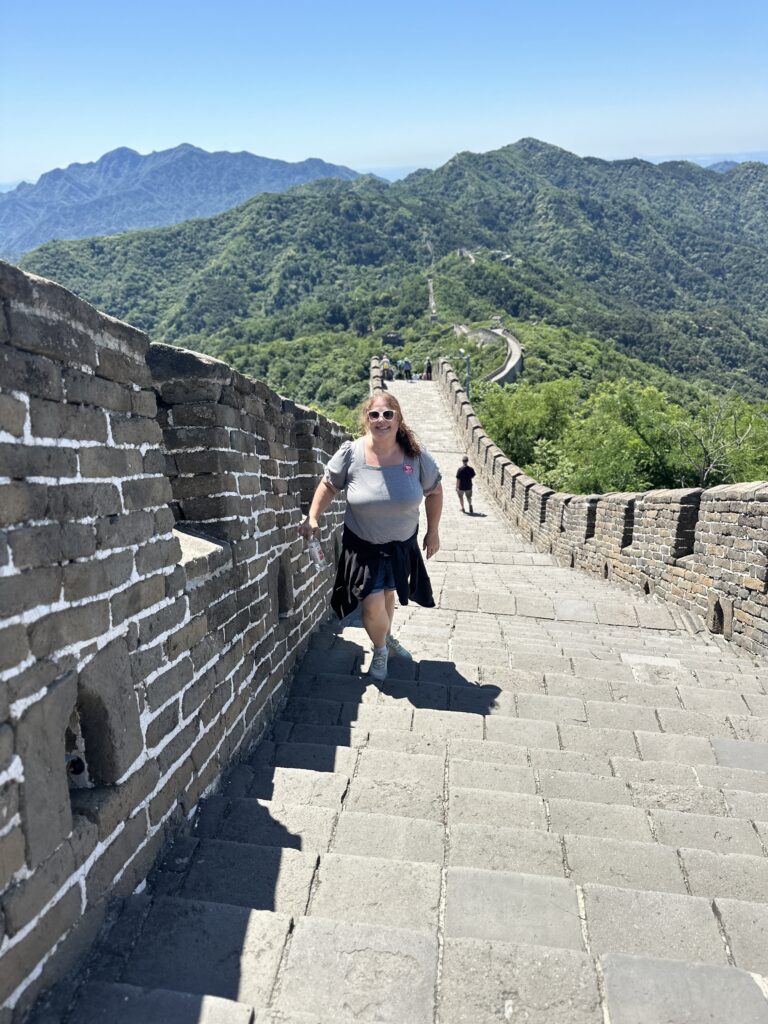 A person is walking up a stone staircase on the Great Wall of China. The wall stretches into the distance, surrounded by lush green mountains under a clear blue sky. The person is wearing sunglasses, a gray shirt, and shorts, and is holding a water bottle.