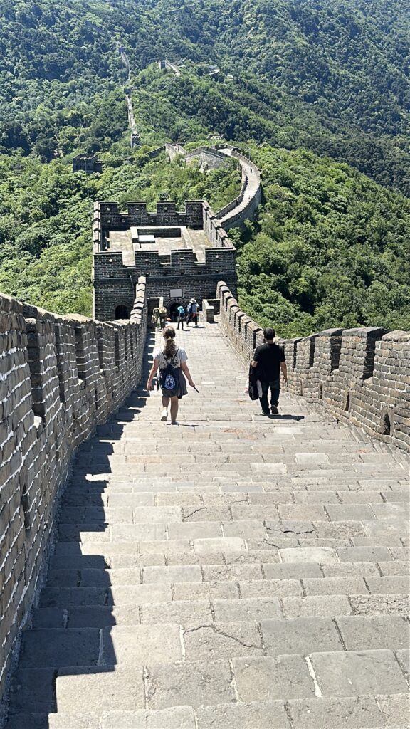 A view of the Great Wall of China, showing a long stretch of stone steps and walls extending into the distance. Several people are walking along the wall, surrounded by lush green hills and forests. The sky is clear, and the scene is brightly lit by sunlight.