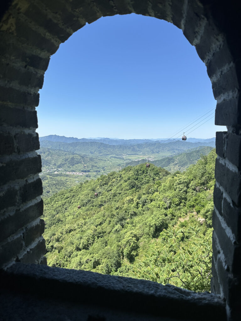 A view through a brick archway shows a lush, green mountainous landscape under a clear blue sky. In the distance, a cable car is visible, suspended on wires above the hills.