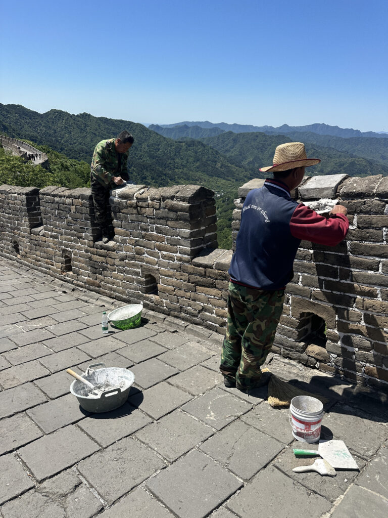 Two workers are repairing a section of the Great Wall of China. One is standing on the wall, applying mortar to the bricks, while the other is standing on the walkway, also working on the wall. They are surrounded by tools and materials, including buckets and brushes. The background features a scenic view of green mountains under a clear blue sky.