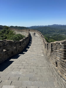 A section of the Great Wall of China under a clear blue sky. The stone pathway is flanked by brick walls, with two people walking along it. Lush green hills and distant mountains are visible in the background.