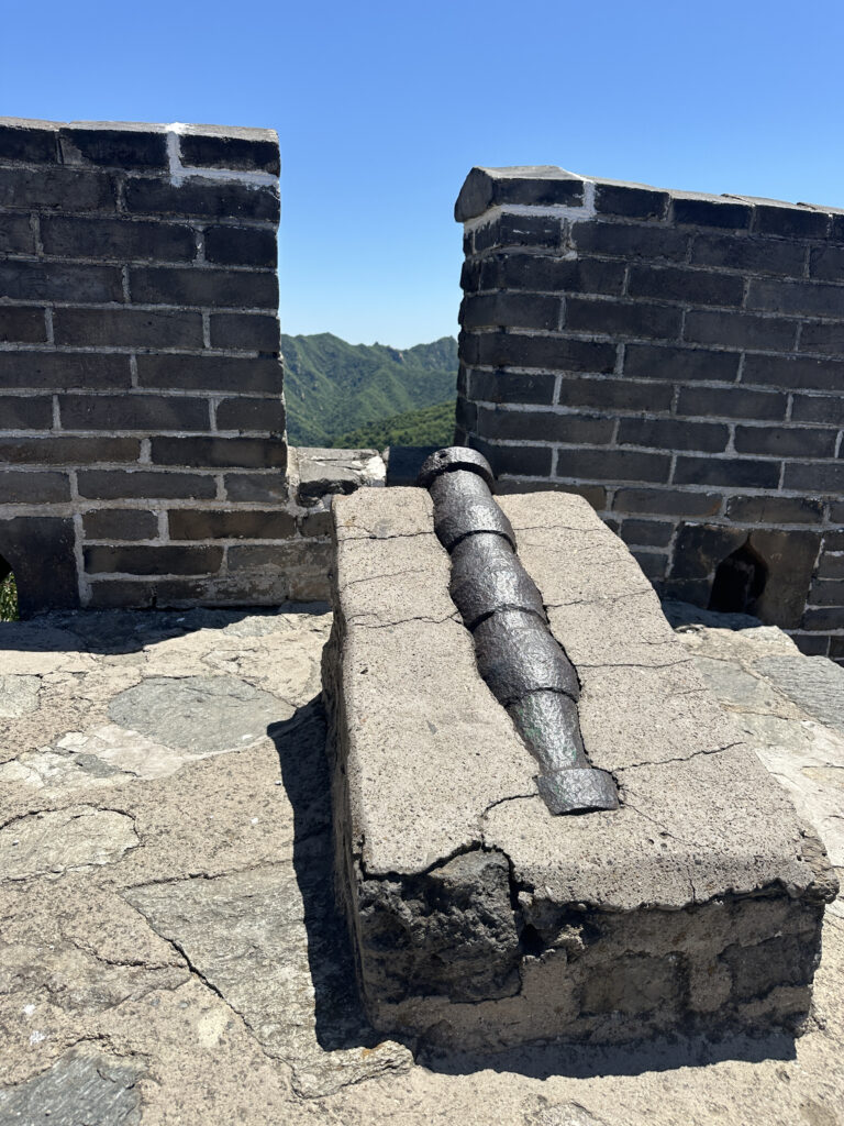 A section of the Great Wall of China with a cannon placed on a stone platform. The wall is made of gray bricks, and there is a gap between two sections of the wall. In the background, green hills are visible under a clear blue sky.