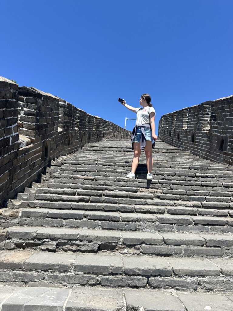 A person is standing on a stone staircase of the Great Wall of China, taking a selfie with a smartphone. The sky is clear and blue, and the person is wearing casual clothing with a jacket tied around their waist. The stone walls of the Great Wall are visible on either side.