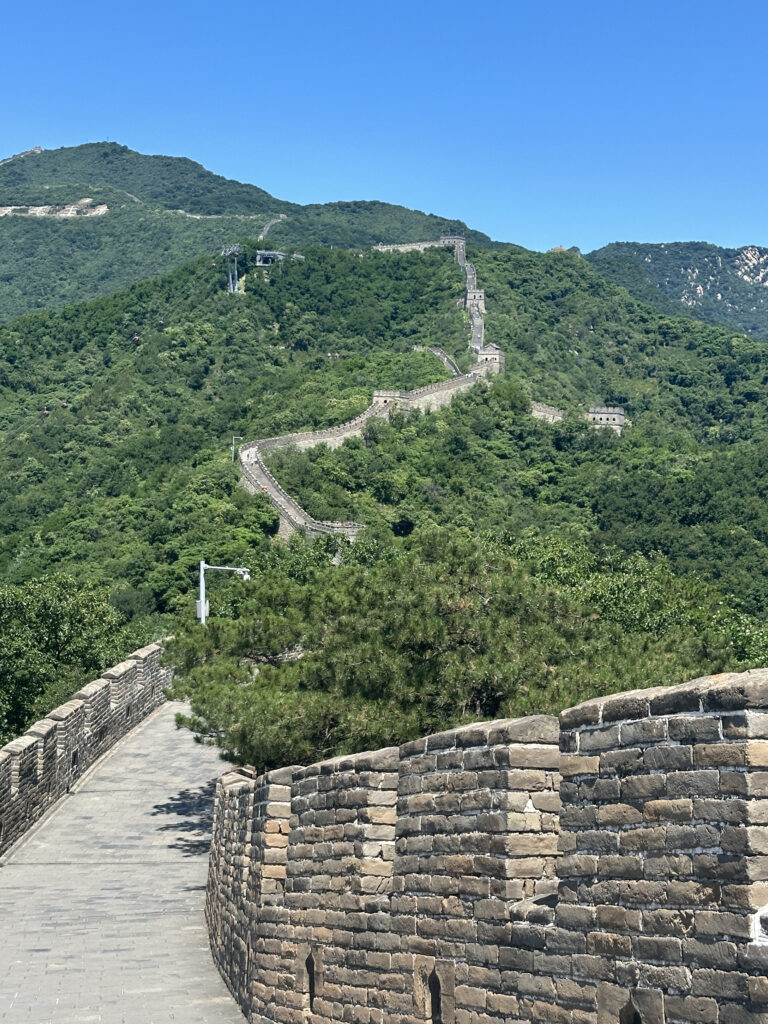 The image shows a section of the Great Wall of China winding over lush green hills under a clear blue sky. The stone wall features battlements and a pathway, with trees and vegetation surrounding the area.