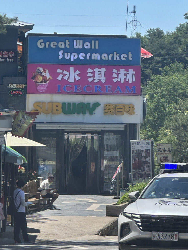The image shows the entrance to a building with multiple signs. At the top, there is a sign for "Great Wall Supermarket." Below it, there is a pink sign with Chinese characters and the word "ICE CREAM." Another sign below reads "SUBWAY" with additional Chinese characters. A police car is parked in the foreground on the right, and a few people are visible near the entrance. There are trees and a power line in the background.