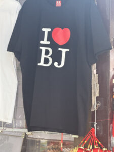 A black T-shirt is hanging on display. It features the text "I ❤️ BJ" in large white letters with a red heart symbol. The shirt is part of a market or store setting, with other items visible in the background.