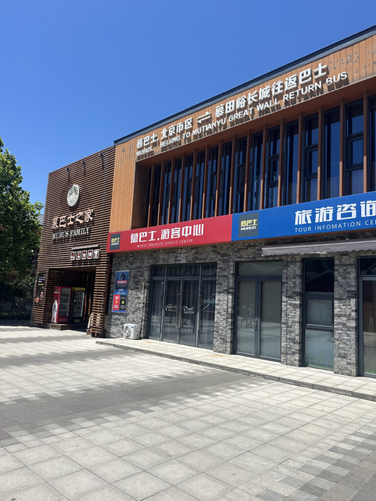 The image shows a building with signs in both English and Chinese. The signs indicate it is a service center for "Mubus," offering a return bus service from Beijing to the Mutianyu Great Wall. The building has a modern design with a combination of wood and stone elements. There are large windows and a vending machine visible near the entrance. The sky is clear and blue, and there are trees on the left side of the image.