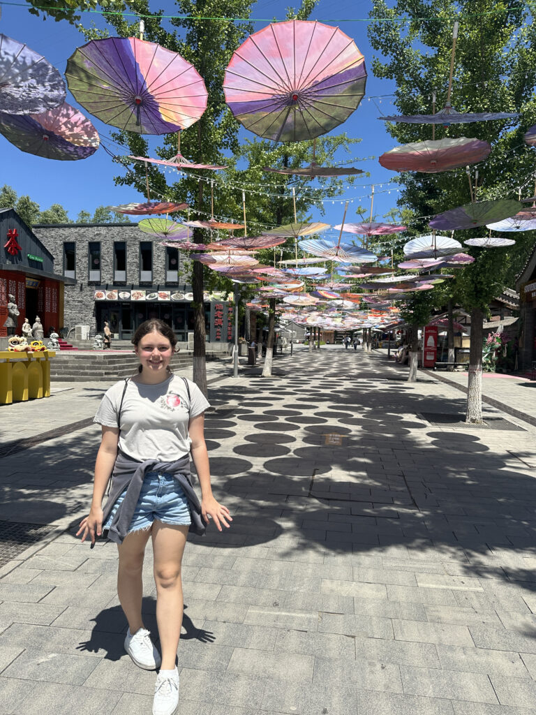 A person is standing on a paved street lined with trees. Above, colorful umbrellas are suspended, creating a decorative canopy. The street is in a sunny area with buildings and shops in the background. The person is wearing a light-colored t-shirt, denim shorts, and white sneakers, with a jacket tied around their waist.