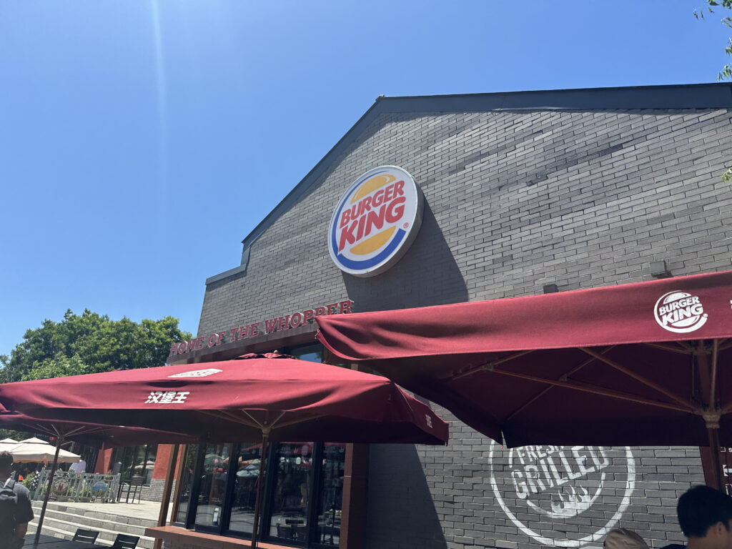 The image shows the exterior of a Burger King restaurant. The building is made of gray bricks and features the Burger King logo prominently on the wall. Below the logo, the phrase "Home of the Whopper" is visible. There are maroon umbrellas with the Burger King logo providing shade for outdoor seating. The sky is clear and blue, and there are some trees in the background.